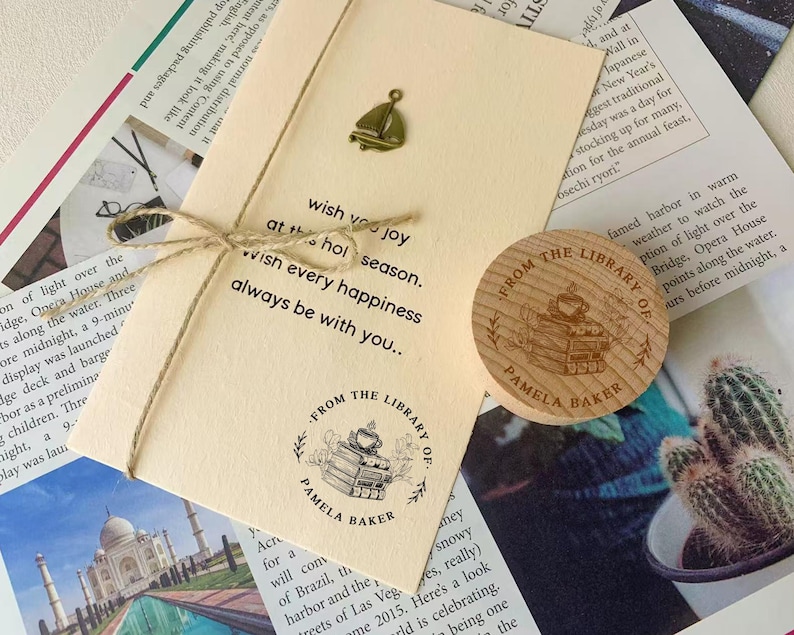 May include: A beige card tied with twine, featuring a sailboat charm and the text "wish you joy...wish every happiness...always be with you." A wooden coaster with "FROM THE LIBRARY OF PAMELA BAKER" is also present.
