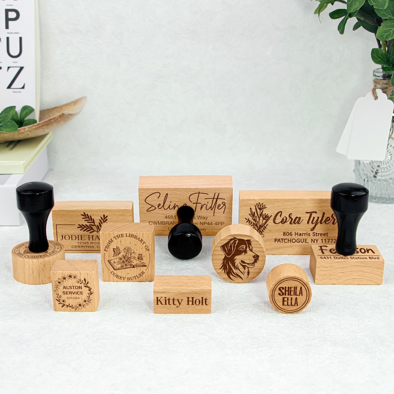May include: A set of nine wooden stamps with various designs, including a dog, a book, a wreath, and text. The stamps are made of natural wood and have a rustic look. The text on the stamps includes names, addresses, and phrases.