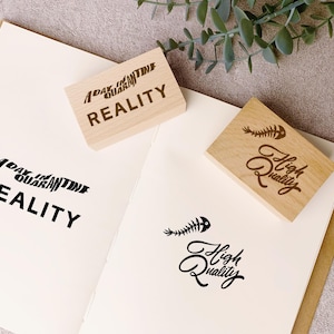 May include: Two wooden stamps with text and a fishbone graphic are on a white paper. One stamp reads "A Day in Quarantine Reality", and the other says "High Quality". The stamped images are in black.