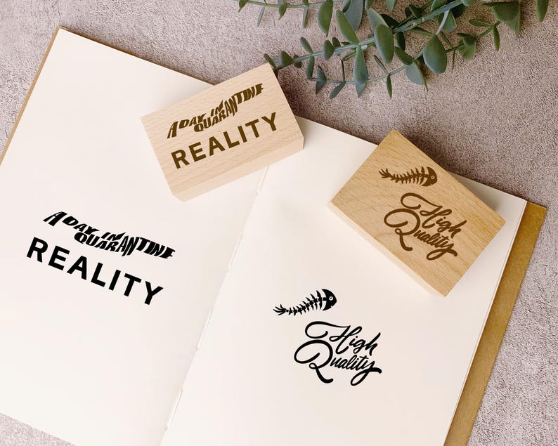 May include: Two wooden stamps with text and a fishbone graphic are on a white paper. One stamp reads "A Day in Quarantine Reality", and the other says "High Quality". The stamped images are in black.