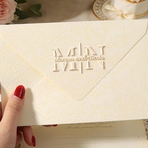 Peut inclure: Enveloppe ivoire avec texte en relief &laquo; Morgan and Nicola &raquo;. L'enveloppe est tenue par une main avec du vernis &agrave; ongles rouge. Le fond comprend des fleurs, un g&acirc;teau et un verre.
