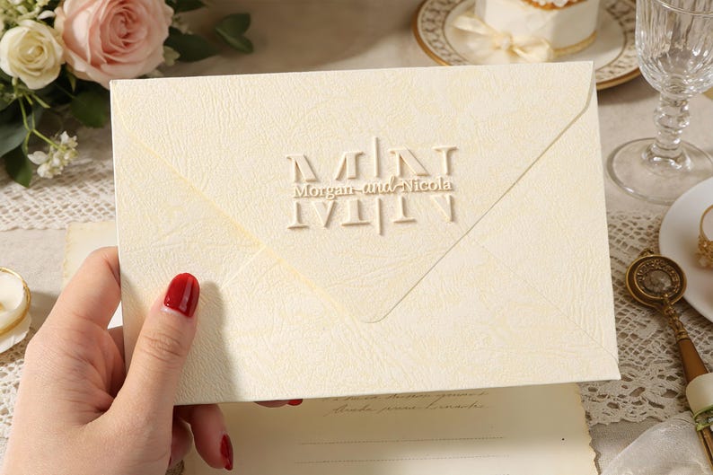 Peut inclure: Enveloppe ivoire avec texte en relief &laquo; Morgan and Nicola &raquo;. L'enveloppe est tenue par une main avec du vernis &agrave; ongles rouge. Le fond comprend des fleurs, un g&acirc;teau et un verre.