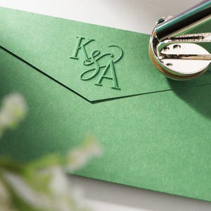 Peut inclure: Une enveloppe verte avec les initiales "K&A" en relief est pr&eacute;sent&eacute;e avec une machine &agrave; imprimer argent&eacute;e et dor&eacute;e. L'enveloppe est de couleur vert sourd. La machine &agrave; imprimer est partiellement visible en haut &agrave; droite.