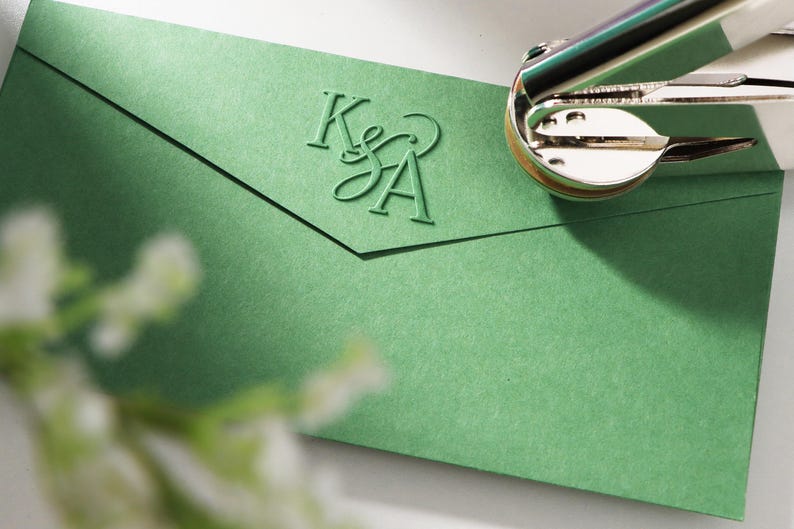 Peut inclure: Une enveloppe verte avec les initiales "K&A" en relief est pr&eacute;sent&eacute;e avec une machine &agrave; imprimer argent&eacute;e et dor&eacute;e. L'enveloppe est de couleur vert sourd. La machine &agrave; imprimer est partiellement visible en haut &agrave; droite.