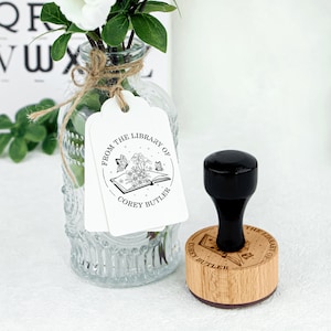 May include: A wooden stamp with a black handle. The stamp features a circular design with the text "From the Library of Corey Butler" and an illustration of an open book with butterflies and flowers.