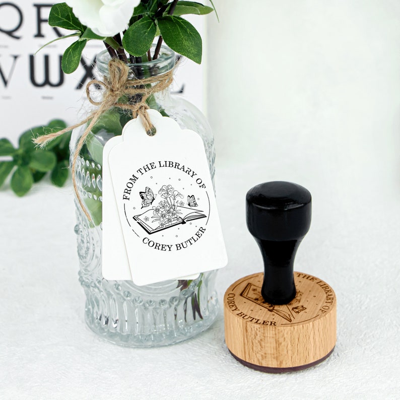 May include: A wooden stamp with a black handle. The stamp features a circular design with the text "From the Library of Corey Butler" and an illustration of an open book with butterflies and flowers.