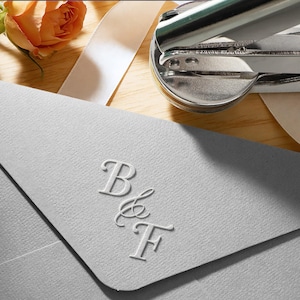 Puede incluir: Un sobre gris claro con las iniciales en relieve "B & F". Un sello plateado y rosas naranjas con una cinta blanca están en el fondo. El sobre está sobre una superficie de madera.