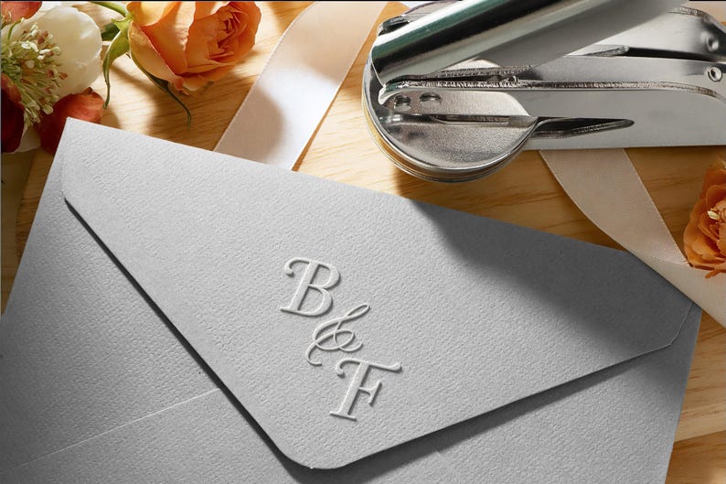 Peut inclure: Une enveloppe gris clair avec les initiales en relief "B & F". Un sceau argent&eacute; et des roses orange avec un ruban blanc sont en arri&egrave;re-plan. L'enveloppe est sur une surface en bois.
