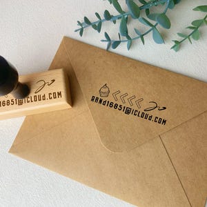 May include: A wooden stamp with a black handle and the text "RAND16851@ICLOUD.COM" and a cupcake icon. The stamp is next to a brown envelope with the same text and cupcake icon. Green leaves are in the background.