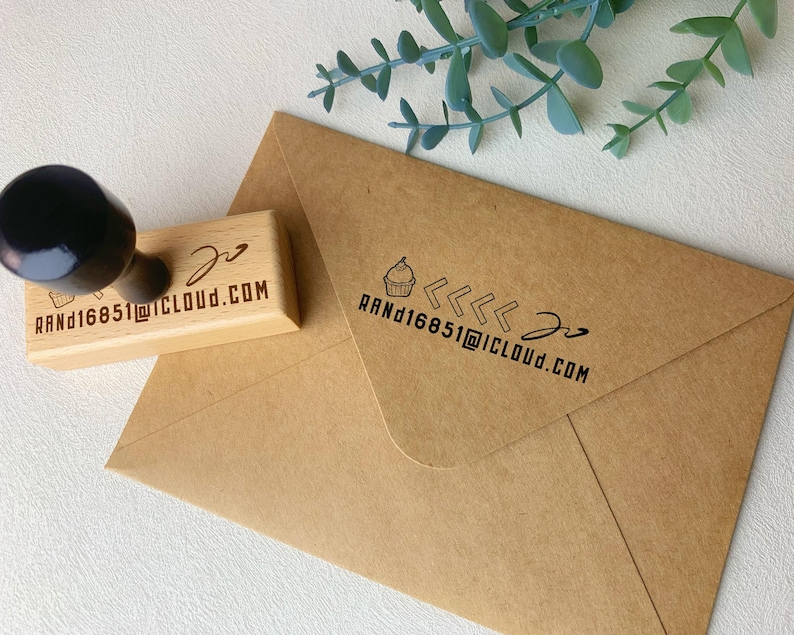 May include: A wooden stamp with a black handle and the text "RAND16851@ICLOUD.COM" and a cupcake icon. The stamp is next to a brown envelope with the same text and cupcake icon. Green leaves are in the background.
