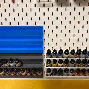 Könnte beinhalten: Eine weiße Lochwand mit zwei Reihen von Farbflaschen, einer Reihe von grauen Farbflaschenhaltern und einer Reihe von blauen Farbflaschenhaltern.