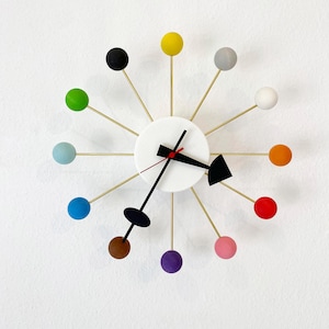Könnte beinhalten: Eine moderne Wanduhr mit weißem Zifferblatt und farbenfrohen, kugelförmigen Akzenten. Die Uhr hat goldfarbene Speichen, die sich von der Mitte aus erstrecken und jeweils in einer andersfarbigen Kugel enden. Die Zeiger sind schwarz mit einem roten Sekundenzeiger.