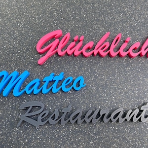 Könnte beinhalten: Drei dimensionale, kursive Schriftbuchstaben, die "Glücklich Matteo Restaurant" in Pink, Blau und Schwarz buchstabieren.