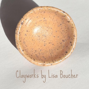 May include: A small, round, speckled ceramic bowl in a warm peach color. The bowl has a slightly irregular shape and a glossy finish. The text "Clayworks by Lisa Boucher" is written in a cursive font below the bowl.