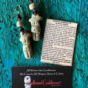 May include: Two dangle earrings featuring light gray clay female figures suspended by dark brown leather cords and small round clay beads. A white informational card with black text and a black card with white text are included in the display.