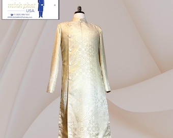 Op de afbeelding: Een crèmekleurige Ao Dai, een traditioneel Vietnamees kledingstuk, wordt op een mannequin getoond. De tuniek met lange mouwen heeft een mandarijnkraag, decoratieve knopen en een ingewikkeld bloemmotief. Het kledingstuk wordt gepresenteerd tegen een neutrale achtergrond.