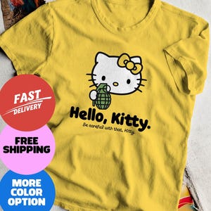 May include: Yellow t-shirt featuring a graphic of Hello Kitty holding a grenade. The text "Hello, Kitty. Be careful with that, Kitty" is printed below the graphic. The shirt is a crew neck style.