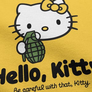 May include: Yellow graphic tee featuring a white cartoon cat with a yellow bow holding a green grenade. The text "Hello, Kitty" is printed in black, with the phrase "Be careful with that, Kitty" below.