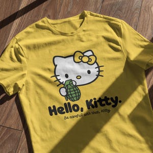 May include: Yellow t-shirt featuring a graphic of Hello Kitty holding a green grenade. The design includes the text "Hello, Kitty." and "Be careful with that, Kitty."