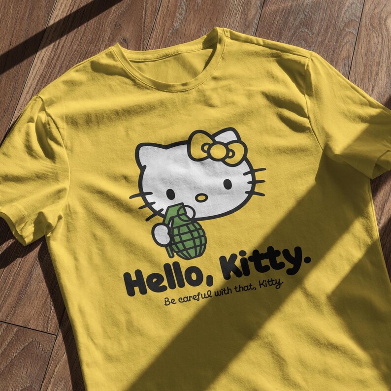 May include: Yellow t-shirt featuring a graphic of Hello Kitty holding a green grenade. The design includes the text "Hello, Kitty." and "Be careful with that, Kitty."