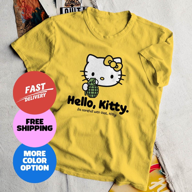 May include: Yellow t-shirt featuring a graphic of Hello Kitty holding a grenade. The text "Hello, Kitty. Be careful with that, Kitty" is printed below the graphic. The shirt is a crew neck style.