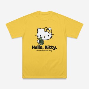 May include: Yellow t-shirt featuring a graphic of Hello Kitty holding a grenade. The text "Hello, Kitty." is printed below the graphic, with the phrase "Be careful with that, Kitty." underneath.