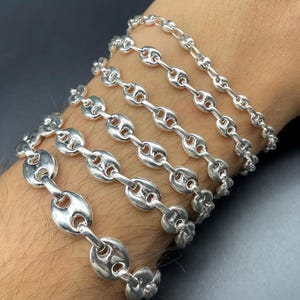 May include: Multiple silver chain bracelets with a coffee bean link design are stacked on a wrist. The bracelets vary in size, with the largest links at the bottom and the smallest at the top. The background is a dark grey.