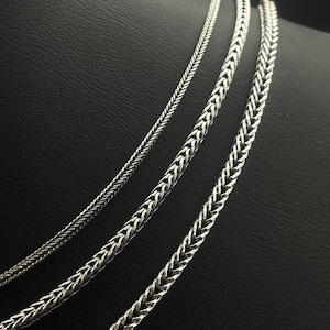 May include: Three silver-colored chain necklaces with different woven patterns are displayed against a black background. The chains vary in thickness, showcasing intricate designs. The necklaces are likely for fashion or jewelry.