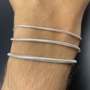 Puede incluir: Tres pulseras plateadas de cadena de serpiente se llevan en la muñeca de una persona. Las pulseras tienen un aspecto texturizado y tejido y se muestran sobre un fondo oscuro. Las pulseras son sencillas y elegantes.
