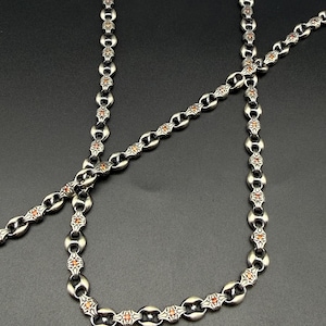 May include: A silver-toned metal chain necklace featuring repeating oval links with intricate engraved detailing and small, embedded orange stones. The chain has a polished finish and a consistent pattern throughout its entire length.