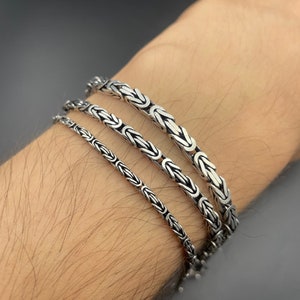 May include: Three silver chain bracelets of varying widths, stacked on a person's wrist.