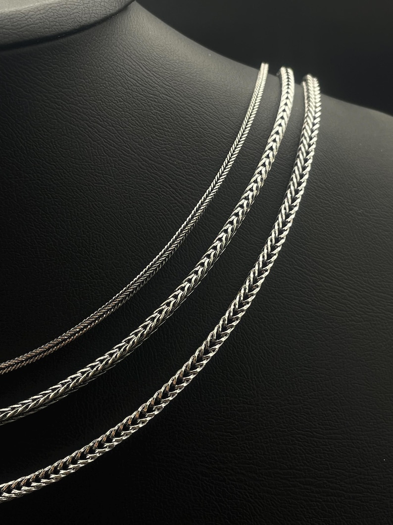 May include: Three silver-colored chain necklaces with different woven patterns are displayed against a black background. The chains vary in thickness, showcasing intricate designs. The necklaces are likely for fashion or jewelry.