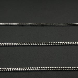 May include: Close-up of three silver-colored chain necklaces with a woven or braided design. The chains are displayed against a dark background, highlighting their texture and metallic sheen. The chains are of varying thicknesses.