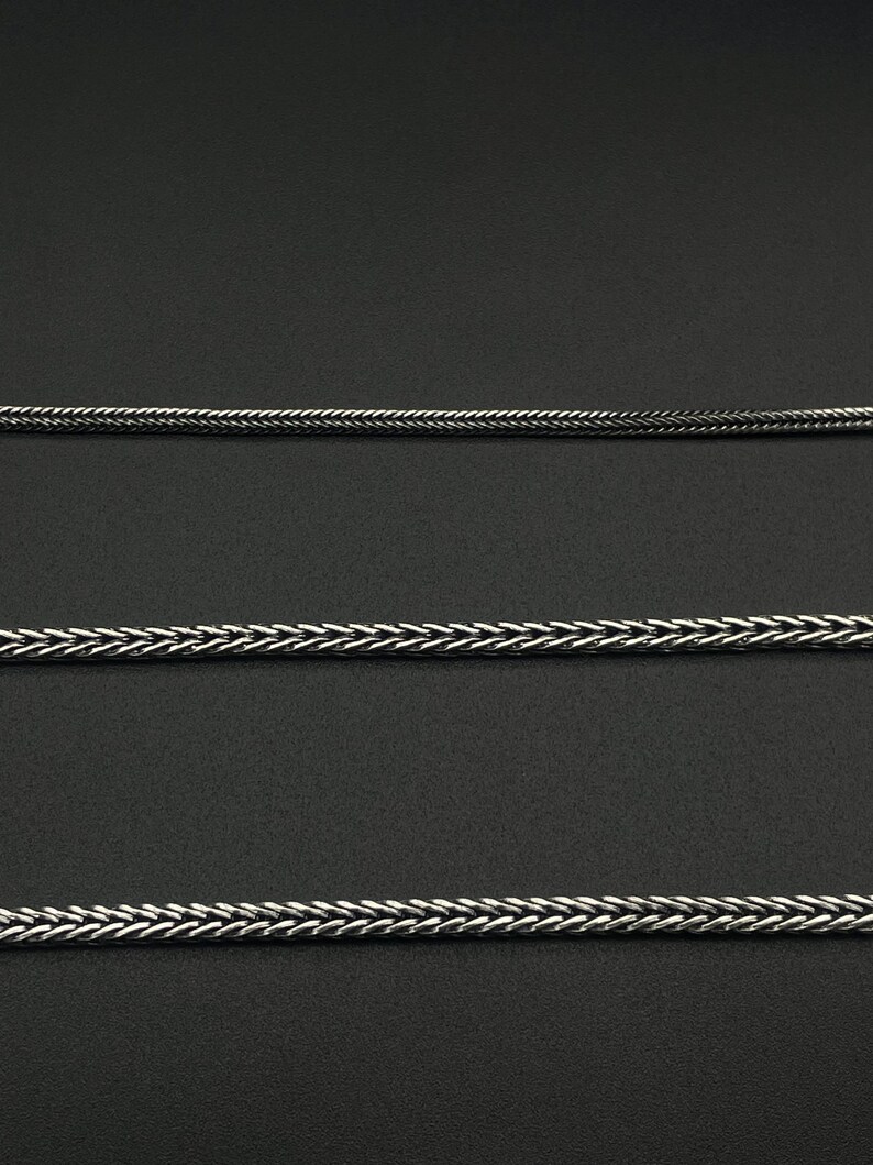 May include: Close-up of three silver-colored chain necklaces with a woven or braided design. The chains are displayed against a dark background, highlighting their texture and metallic sheen. The chains are of varying thicknesses.
