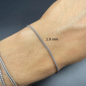May include: A silver bracelet with a woven chain design, 1.8 mm thick, is shown on a wrist. The bracelet is a bright silver color and appears to be made of metal. The background is a dark gray color.