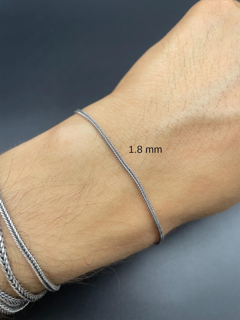 May include: A silver bracelet with a woven chain design, 1.8 mm thick, is shown on a wrist. The bracelet is a bright silver color and appears to be made of metal. The background is a dark gray color.
