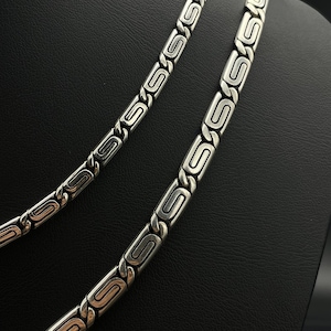 Puede incluir: Dos collares plateados con un dise&ntilde;o geom&eacute;trico repetitivo. Los collares se exhiben sobre una superficie negra, mostrando sus intrincados eslabones y acabado pulido. El dise&ntilde;o presenta una serie de formas de S estilizadas y conectadas.