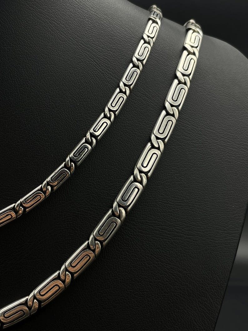 Puede incluir: Dos collares plateados con un dise&ntilde;o geom&eacute;trico repetitivo. Los collares se exhiben sobre una superficie negra, mostrando sus intrincados eslabones y acabado pulido. El dise&ntilde;o presenta una serie de formas de S estilizadas y conectadas.