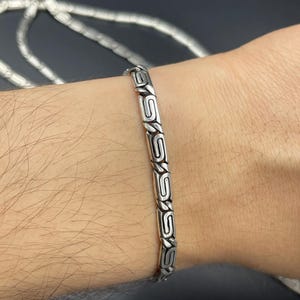 Puede incluir: Se muestra una pulsera de plata con un dise&ntilde;o geom&eacute;trico repetitivo en la mu&ntilde;eca de una persona. La pulsera est&aacute; hecha de segmentos rectangulares unidos con un patr&oacute;n grabado. El fondo es oscuro y otra cadena es visible en el fondo.