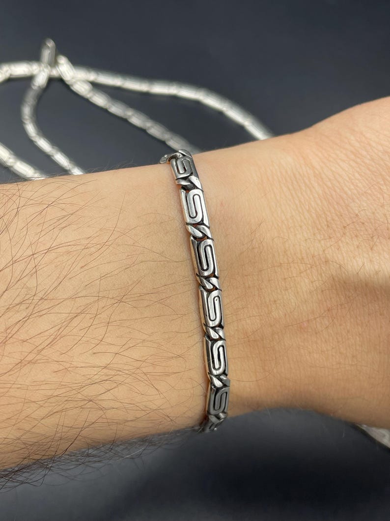 Puede incluir: Se muestra una pulsera de plata con un dise&ntilde;o geom&eacute;trico repetitivo en la mu&ntilde;eca de una persona. La pulsera est&aacute; hecha de segmentos rectangulares unidos con un patr&oacute;n grabado. El fondo es oscuro y otra cadena es visible en el fondo.