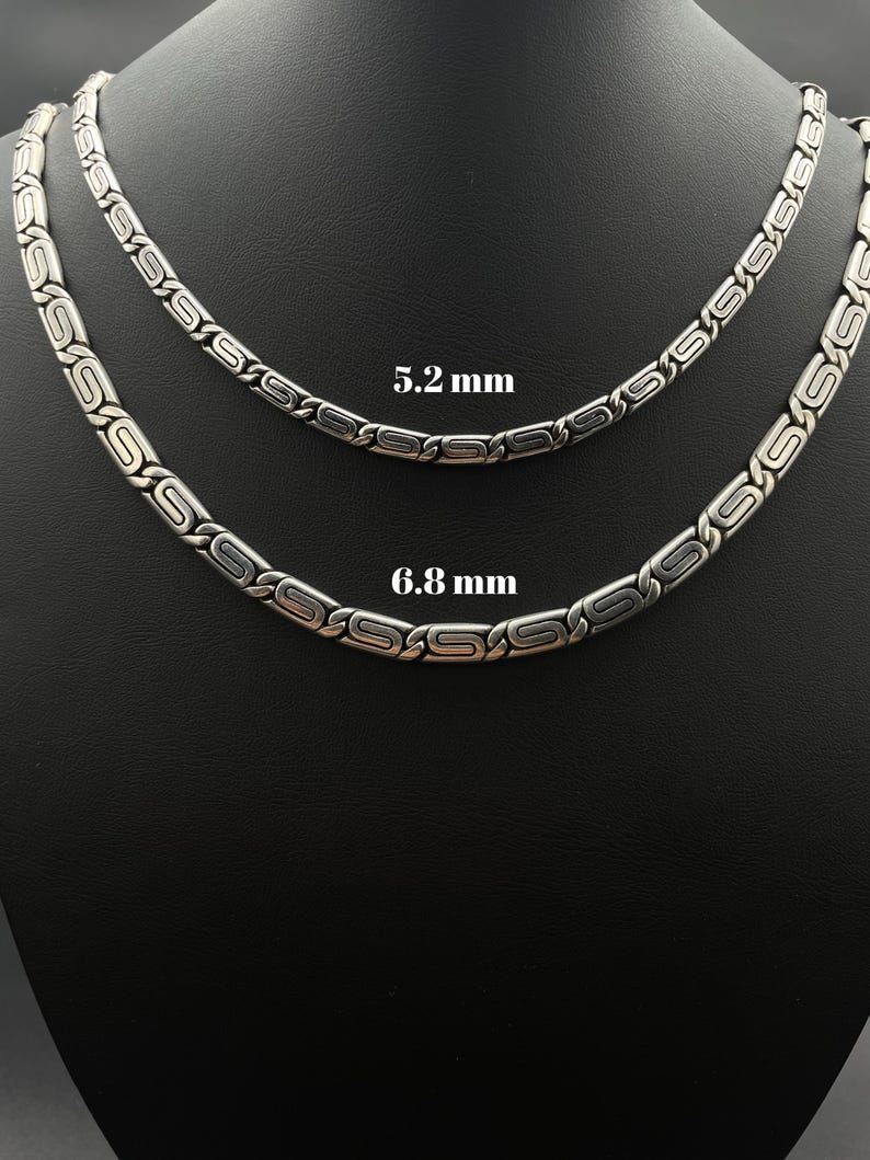 Puede incluir: Dos collares plateados con un dise&ntilde;o repetitivo de &oacute;valos y barras, presentados sobre un fondo negro. Los collares miden 5,2 mm y 6,8 mm de ancho. La joyer&iacute;a se presenta sobre un busto de maniqu&iacute;.