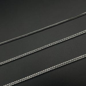 May include: Three silver-colored wheat chain necklaces of varying thicknesses are displayed against a dark background. The chains have a woven design, creating a textured appearance. The necklaces are likely jewelry.