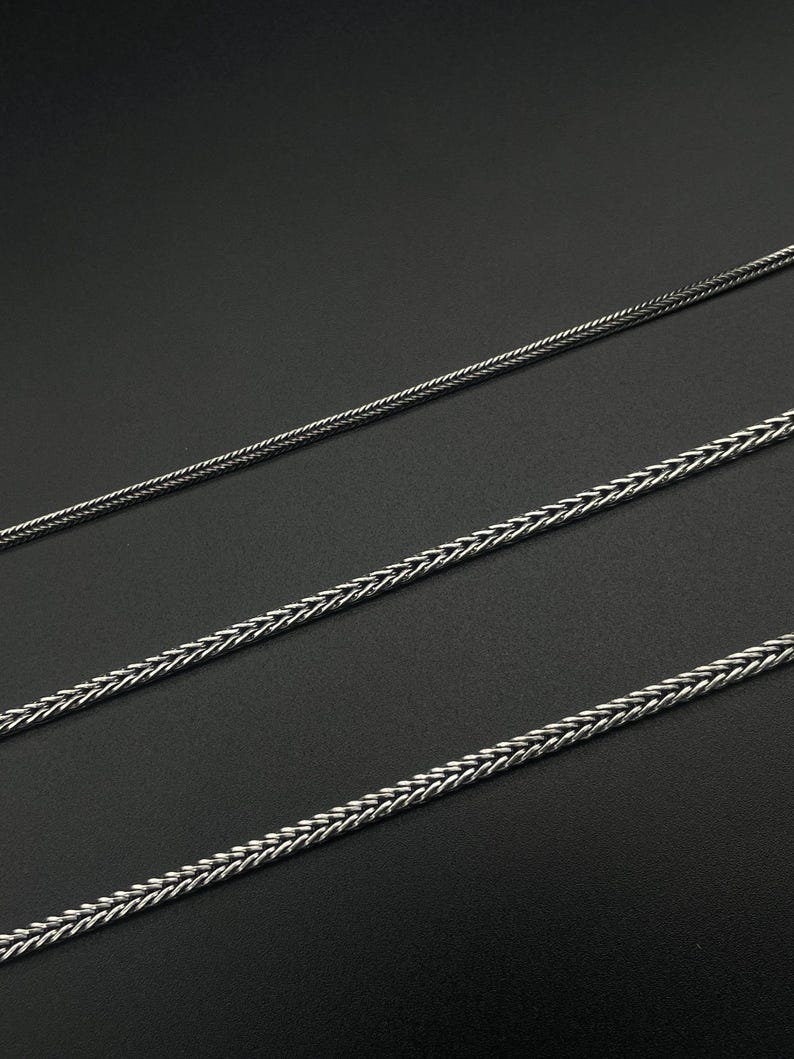 May include: Three silver-colored wheat chain necklaces of varying thicknesses are displayed against a dark background. The chains have a woven design, creating a textured appearance. The necklaces are likely jewelry.