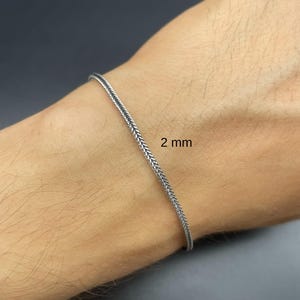 May include: A silver-colored bracelet with a woven design, 2 mm wide, is shown on a wrist. The bracelet has a textured appearance and a simple, elegant style. The background is a dark gray color.