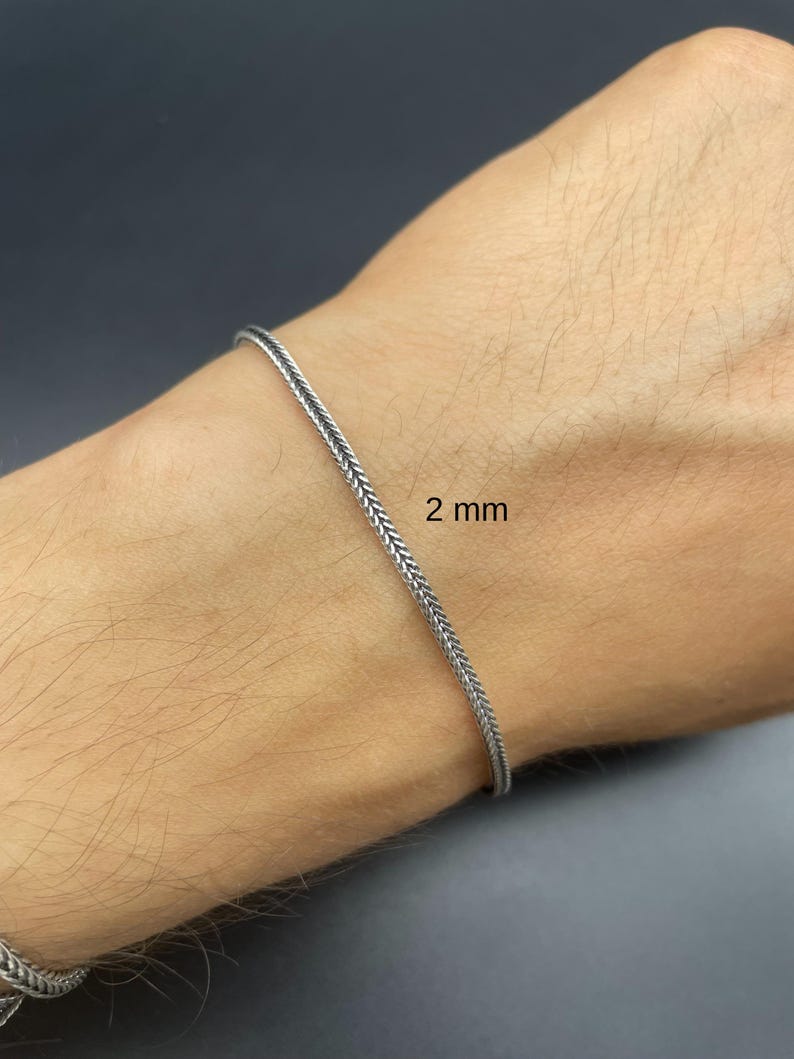 May include: A silver-colored bracelet with a woven design, 2 mm wide, is shown on a wrist. The bracelet has a textured appearance and a simple, elegant style. The background is a dark gray color.