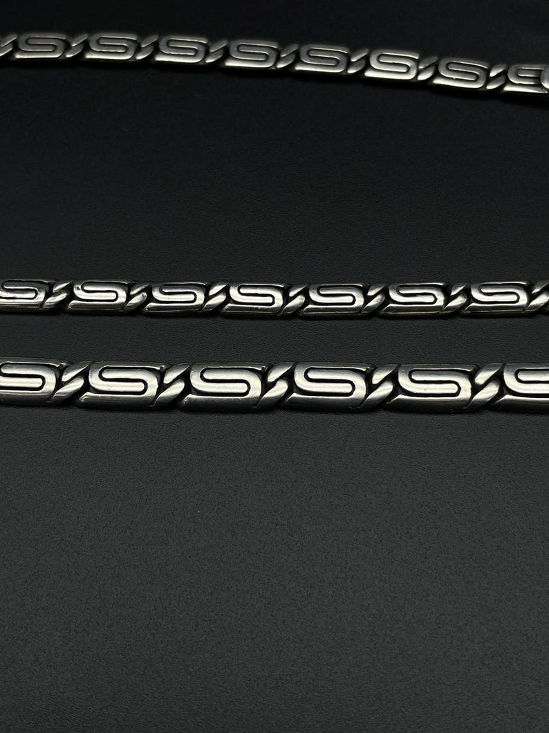 Puede incluir: Primer plano de tres collares de cadena de metal plateado. Las cadenas tienen un dise&ntilde;o de eslabones en forma de S repetidos. El fondo es negro s&oacute;lido.