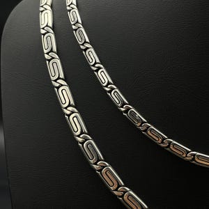 Puede incluir: Dos collares plateados con un dise&ntilde;o geom&eacute;trico repetitivo. Los eslabones son rectangulares con un &oacute;valo central y detalles curvos. Los collares se exhiben sobre un fondo negro, mostrando los detalles de la joyer&iacute;a.