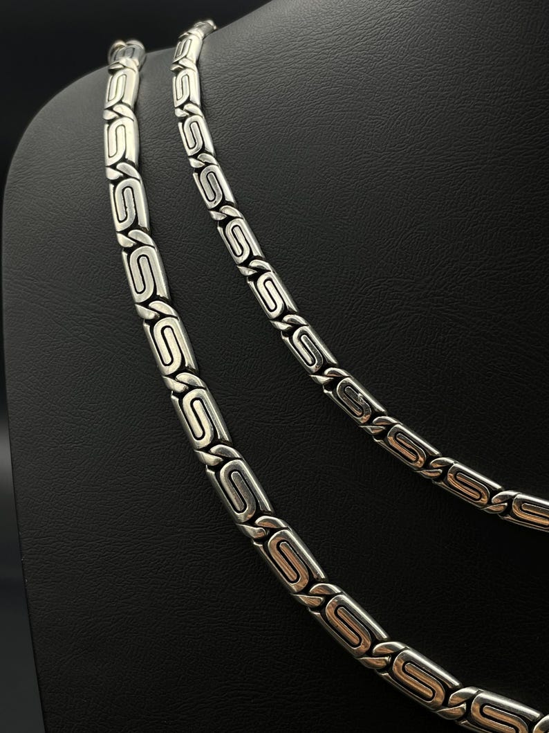 Puede incluir: Dos collares plateados con un dise&ntilde;o geom&eacute;trico repetitivo. Los eslabones son rectangulares con un &oacute;valo central y detalles curvos. Los collares se exhiben sobre un fondo negro, mostrando los detalles de la joyer&iacute;a.