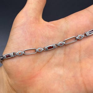 Peut inclure: Un bracelet en chaîne argentée composé de maillons rectangulaires alternant avec des segments métalliques ornés. Ces segments sont sertis de pierres rectangulaires rouge foncé et de petites pierres rondes orange vif en accent.