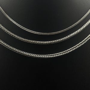 May include: Three silver-colored chain necklaces of varying thicknesses are displayed against a black background. The chains have different woven patterns, with the thickest chain at the bottom and the thinnest at the top. The chains are likely made of metal.